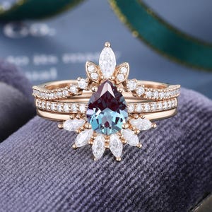 May include: A rose gold ring set featuring a pear-shaped color-changing center stone surrounded by marquise and round-cut white stones. The design includes a curved band with pav&eacute;-set white stones that frames the central pear-shaped gemstone.