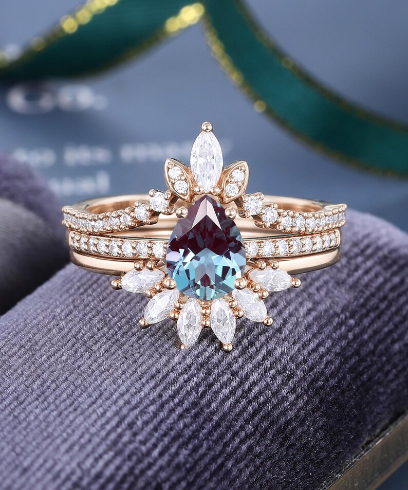 May include: A rose gold ring set featuring a pear-shaped color-changing center stone surrounded by marquise and round-cut white stones. The design includes a curved band with pav&eacute;-set white stones that frames the central pear-shaped gemstone.