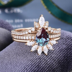 May include: A rose gold ring set featuring a pear-shaped color-changing center stone. The main ring is accented with marquise and round-cut white stones in a sunburst arrangement, while the matching band is paved with small round white stones.