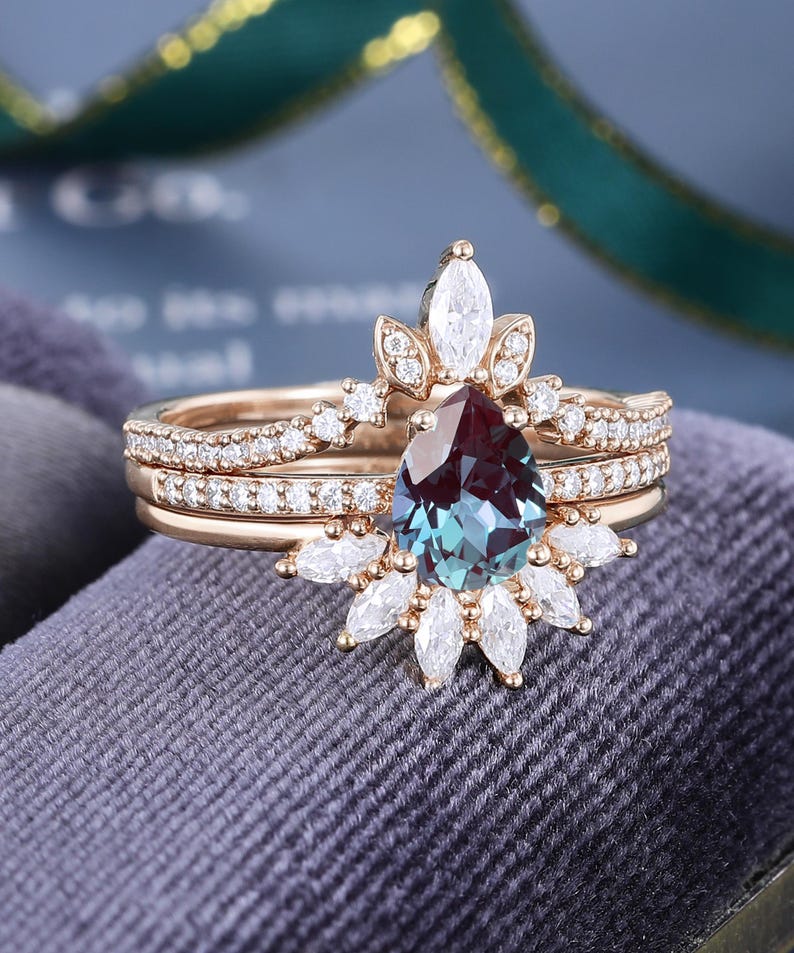 May include: A rose gold ring set featuring a pear-shaped color-changing center stone. The main ring is accented with marquise and round-cut white stones in a sunburst arrangement, while the matching band is paved with small round white stones.