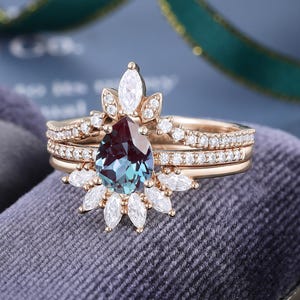 May include: A rose gold ring set featuring a pear-shaped color-changing center stone surrounded by marquise and round-cut white stones. The band is accented with small round white stones, creating a layered look with a crown-like design.