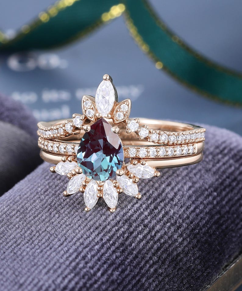 May include: A rose gold ring set featuring a pear-shaped color-changing center stone surrounded by marquise and round-cut white stones. The band is accented with small round white stones, creating a layered look with a crown-like design.