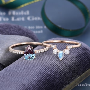 May include: A rose gold ring set featuring a pear-shaped color-changing alexandrite stone on a diamond-encrusted band, paired with a curved band showcasing a central pear-shaped moonstone flanked by two round diamonds.