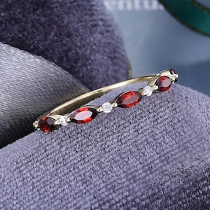 May include: A thin gold band ring featuring an alternating pattern of marquise-cut deep red garnet gemstones and small round clear white stones. The ring is designed with a delicate metal setting that holds each stone securely in a linear arrangement.
