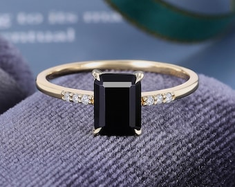 May include: A gold ring featuring a rectangular emerald-cut black gemstone as the center stone, held by four prongs. The thin gold band is accented with three small, round, clear white stones set into the metal on each side of the center stone.