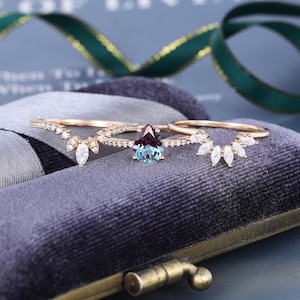 May include: A set of three rose gold rings featuring a central pear-shaped color-changing gemstone flanked by two curved bands. Each band is adorned with small, clear, marquise and round-cut stones set along the thin metal shanks.