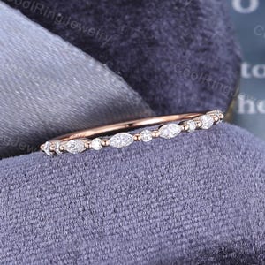 May include: A rose gold band ring featuring an alternating pattern of marquise-cut and round-cut clear stones set along the top half of the band. The design displays a delicate, thin metal profile with a polished finish and clear, sparkling stones.