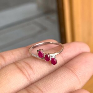 May include: A silver ring featuring three oval-cut, vibrant red gemstones. The gemstones are set in a cluster design, adding a touch of elegance. The ring's band is thin and polished, creating a delicate and refined look.