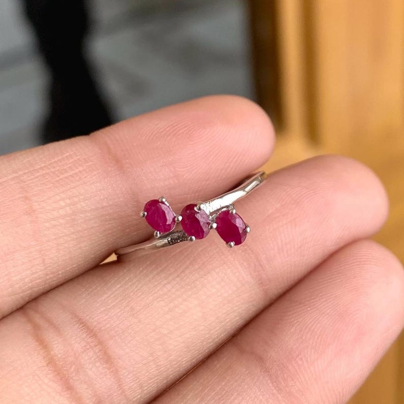 May include: A silver ring featuring three oval-cut, vibrant red gemstones. The gemstones are set in a delicate, curving design, creating a unique and elegant piece of jewelry. The ring is shown on a hand, highlighting its size and style.