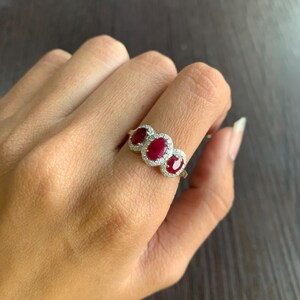 May include: A silver ring featuring three oval-cut, deep red gemstones, each surrounded by a halo of smaller, clear stones. The ring is set against a dark background, highlighting the sparkle of the gems.