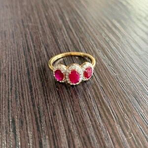 May include: A gold ring featuring three oval-cut, vibrant red gemstones, each surrounded by a halo of smaller, clear stones. The ring's band is a polished gold, creating a classic and elegant design. The ring is set against a dark wood grain surface.