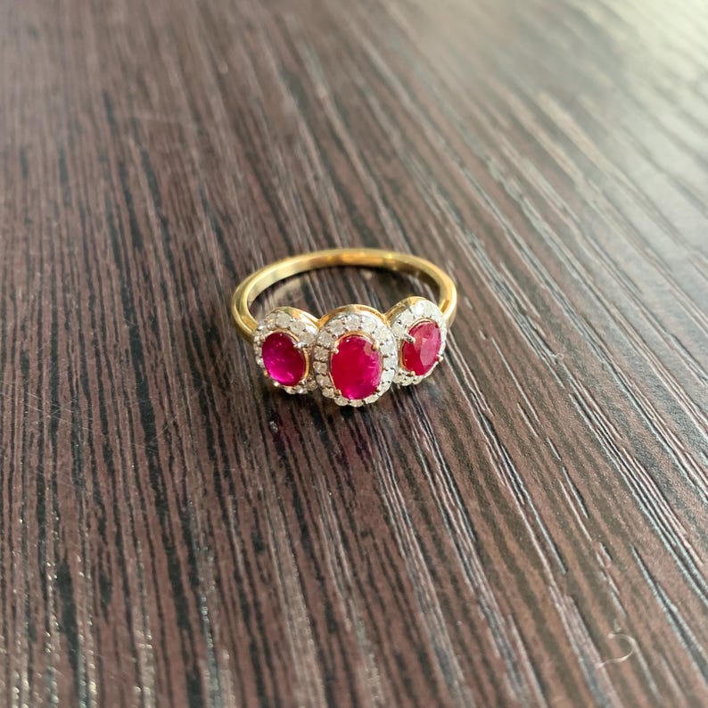 May include: A gold ring featuring three oval-cut, vibrant red gemstones, each surrounded by a halo of smaller, clear stones. The ring's band is a polished gold, creating a classic and elegant design. The ring is set against a dark wood grain surface.
