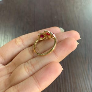 May include: A gold ring with three red gemstones and small white accent stones. The ring is set on a gold band and is displayed on a person's hand.