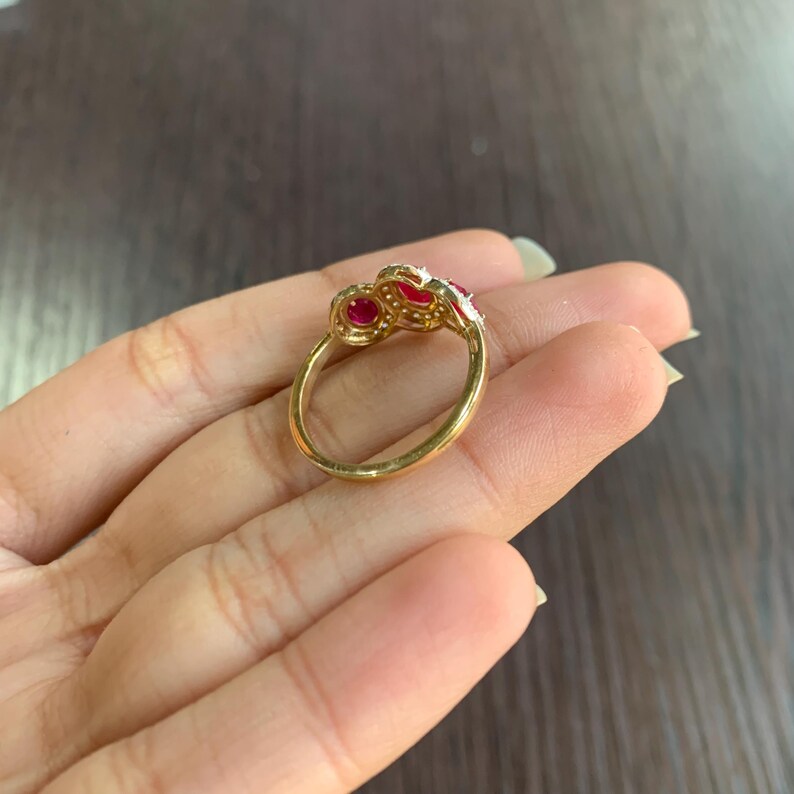 May include: A gold ring with three red gemstones and small white accent stones. The ring is set on a gold band and is displayed on a person's hand.