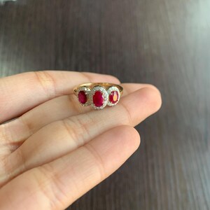 May include: A gold ring featuring three oval-cut, deep red gemstones, each surrounded by a halo of small, clear stones. The ring is set on a slender gold band, showcasing a classic and elegant design.