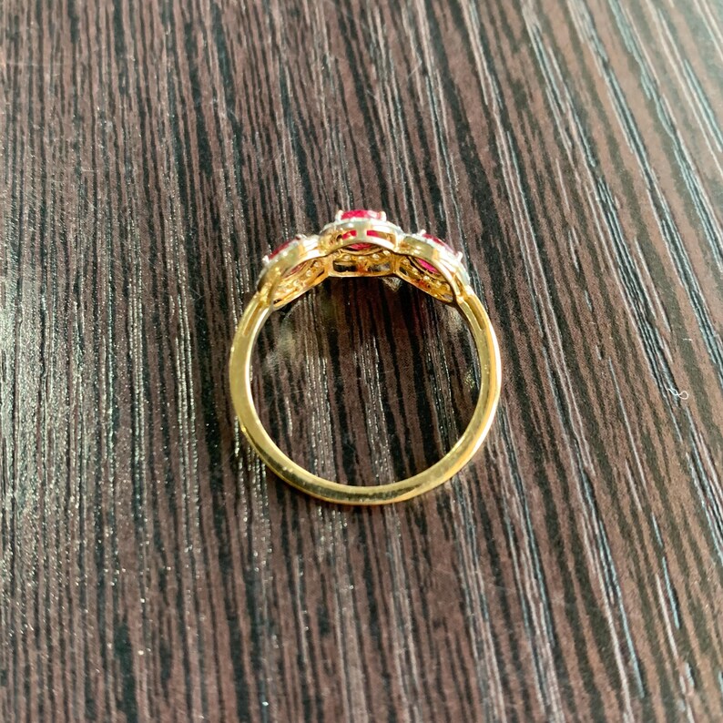 May include: A gold ring with three red gemstones. The ring's band is a simple, polished gold, while the setting around the gemstones has a more ornate, filigree design. The gemstones are a vibrant red color.