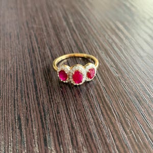 May include: A gold ring featuring three oval-cut, vibrant red gemstones, each surrounded by a halo of smaller, clear stones. The ring's band is a simple, polished gold. The ring is displayed on a dark wood surface.