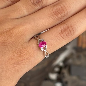 May include: A silver ring with a vivid oval-cut ruby gemstone. The ring has a delicate, intertwined design with small, clear accent stones either side of the ruby. The ring is shown on a finger.