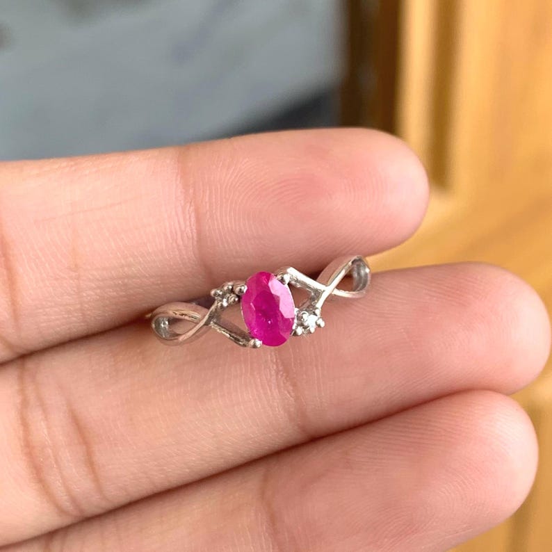 May include: A silver ring with a vivid oval ruby gemstone at its centre. The ring has an intricate design with small accent stones and a delicate, swirling band. The ruby is a deep, rich pink colour.
