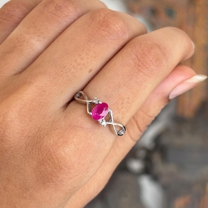 May include: A silver ring with a vivid oval ruby gemstone. The ring has an infinity symbol design on each side of the ruby, with small accent stones. The ring is shown on a hand.
