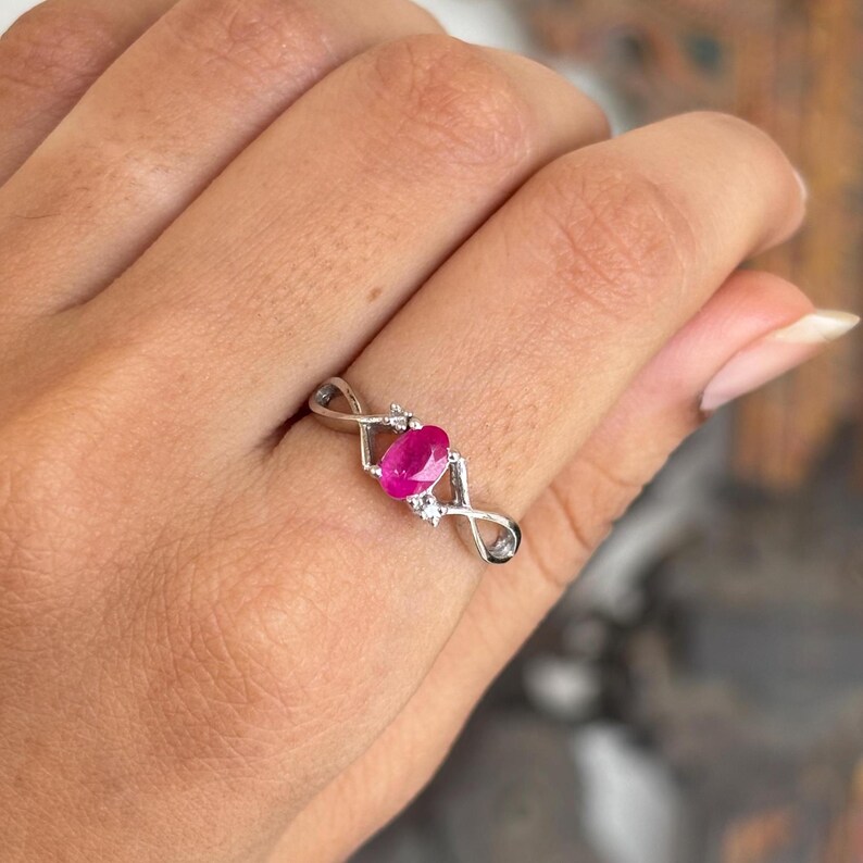 May include: A silver ring with a vivid oval ruby gemstone. The ring has an infinity symbol design on each side of the ruby, with small accent stones. The ring is shown on a hand.