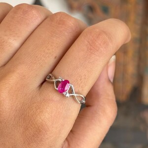 May include: A silver ring with a central oval, vivid red gemstone. The band has an infinity symbol design, with small, clear accent stones either side of the main stone. The ring is displayed on a finger.