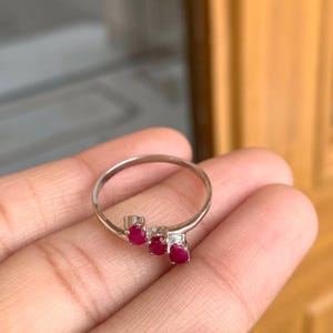 May include: A silver ring featuring three oval-cut, vibrant red gemstones. The ring has a delicate, minimalist design, with the gemstones set in a row. The ring is shown on a hand.
