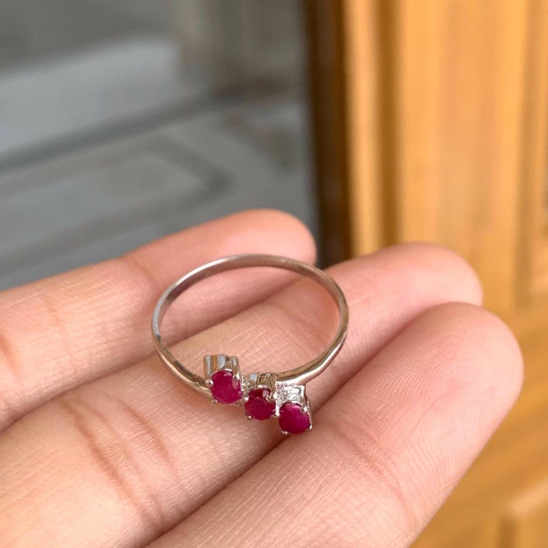 May include: A silver ring featuring three oval-cut, vibrant red gemstones. The ring has a delicate, minimalist design, with the gemstones set in a row. The ring is shown on a hand.