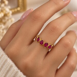 May include: A gold band ring featuring a series of vibrant, teardrop-shaped magenta stones set in a continuous row. The metal has a warm, polished finish, and each stone is securely held within a smooth, rounded bezel setting along the band.