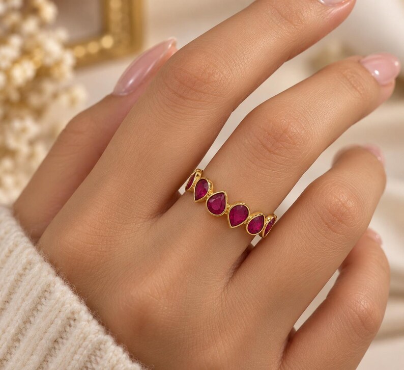 May include: A gold band ring featuring a series of vibrant, teardrop-shaped magenta stones set in a continuous row. The metal has a warm, polished finish, and each stone is securely held within a smooth, rounded bezel setting along the band.
