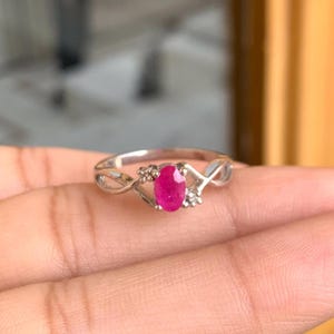 May include: A silver ring featuring a vivid oval ruby gemstone. The ring has an intricate, interwoven pattern on the band, with small accent stones flanking the ruby. The ruby is a deep red hue, set in a delicate silver setting.