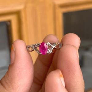 May include: A silver ring with an oval-cut ruby gemstone. The ring has a twisted band design with small accent stones either side of the ruby. The ruby is a deep pink colour. The ring is a piece of jewellery.