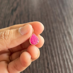 May include: A gold-framed pendant with a bright pink, teardrop-shaped gemstone. The pendant is suspended from a small gold loop. The gemstone's surface is multi-faceted.