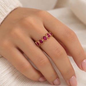May include: A gold band ring featuring a series of five teardrop-shaped, deep pink stones set in a row. The stones are arranged horizontally along the finger, with each stone encased in a smooth gold bezel setting that follows the teardrop shape.