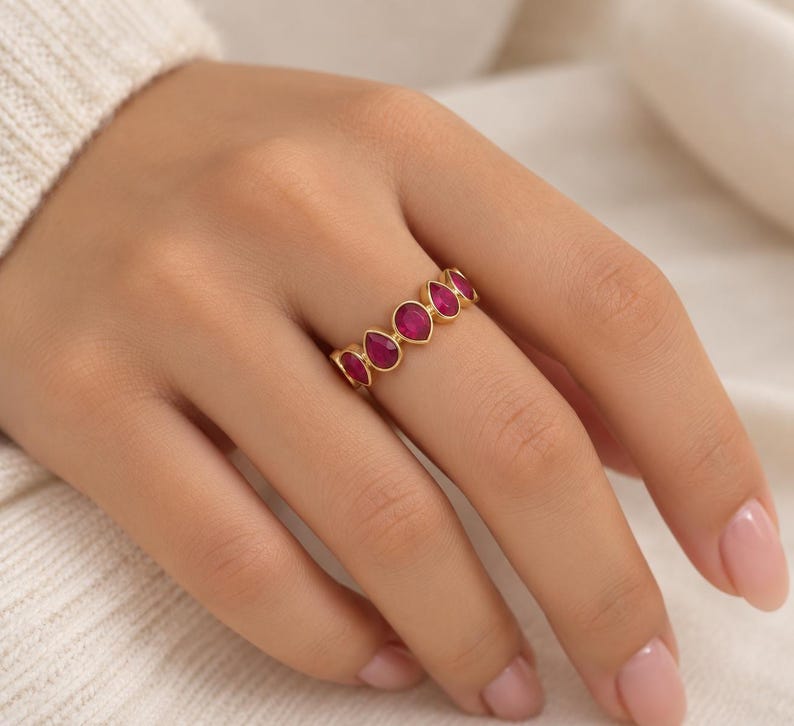 May include: A gold band ring featuring a series of five teardrop-shaped, deep pink stones set in a row. The stones are arranged horizontally along the finger, with each stone encased in a smooth gold bezel setting that follows the teardrop shape.