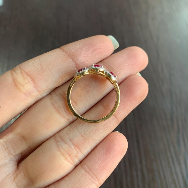 May include: A gold ring featuring three oval-shaped red gemstones, each surrounded by smaller, clear stones. The ring's band is a polished gold color, and the gemstones are set in a decorative pattern. The ring is shown on a hand.