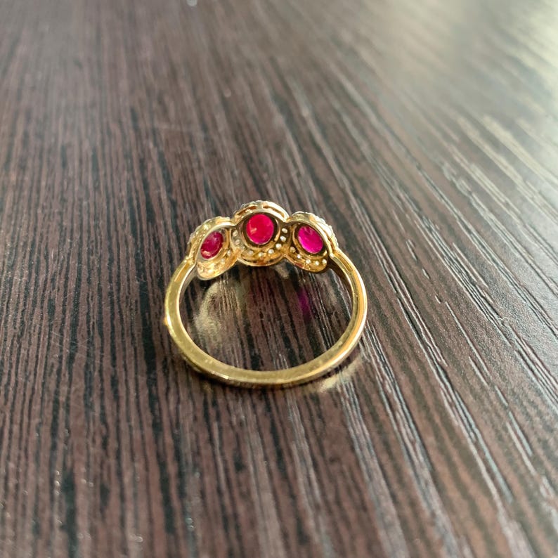 May include: A gold ring featuring three oval-shaped, vibrant red gemstones. The ring's band is a polished gold, and the gemstones are surrounded by smaller, clear stones. The ring is set against a dark wood grain surface.