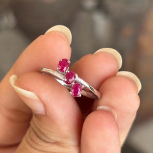 May include: A silver ring featuring three oval-cut, vibrant red gemstones. The ring's band has a delicate, flowing design, with the gemstones set in a row. The ring is held up by a person's fingers.