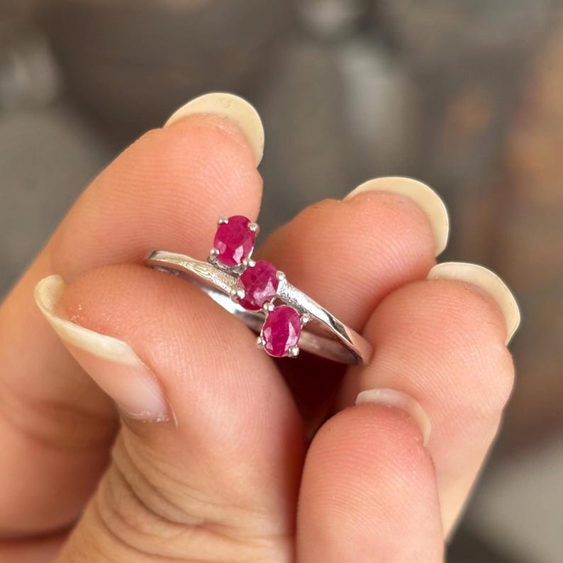 May include: A silver ring featuring three oval-cut, vibrant red gemstones. The ring's band has a delicate, flowing design, with the gemstones set in a row. The ring is held up by a person's fingers.