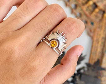 May include: A silver ring featuring a sunburst design with a round, yellow gemstone at its center. The ring's rays extend outward, creating a striking visual effect. The ring is worn on a finger.