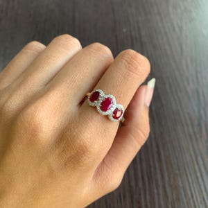 May include: A gold ring featuring three oval-cut, deep red gemstones, each surrounded by a halo of small, clear stones. The ring is set on a hand, showcasing the jewelry's design and color contrast against the skin.