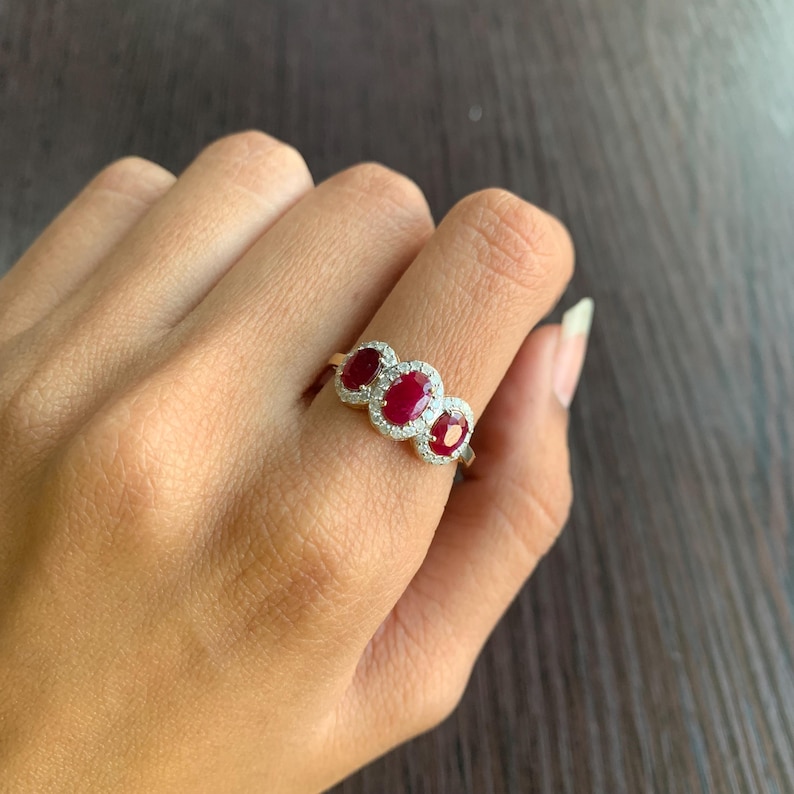 May include: A gold ring featuring three oval-cut, deep red gemstones, each surrounded by a halo of small, clear stones. The ring is set on a hand, showcasing the jewelry's design and color contrast against the skin.