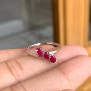 May include: A silver ring featuring three oval-cut, vibrant red gemstones. The gemstones are set in a row, creating a unique and elegant design. The ring is displayed on a hand, showcasing its delicate details.