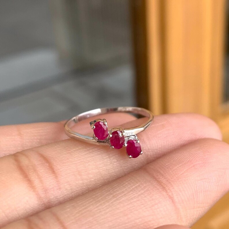May include: A silver ring featuring three oval-cut, vibrant red gemstones. The gemstones are set in a row, creating a unique and elegant design. The ring is displayed on a hand, showcasing its delicate details.