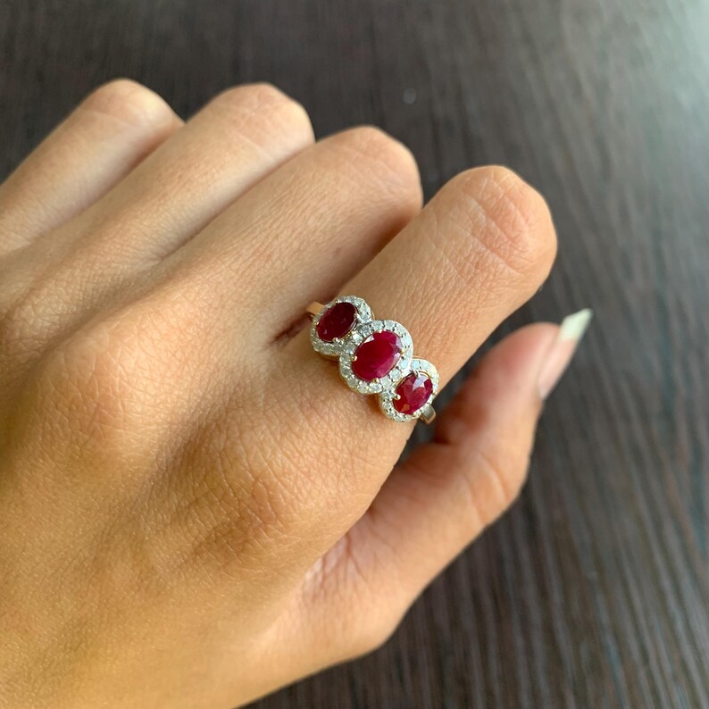 May include: A gold ring featuring three oval-cut rubies, each surrounded by a halo of small, clear gemstones. The rubies are a deep red color, and the ring is set on a finger.