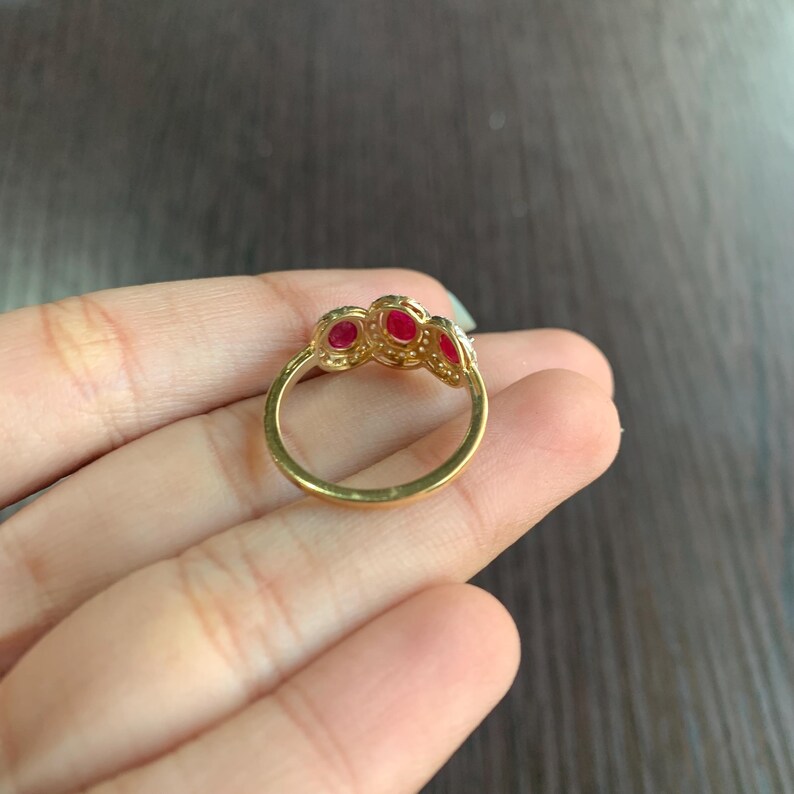 May include: A gold ring featuring three oval-shaped red gemstones. The ring has a delicate band and is set with smaller, clear stones around the red gems. The ring is shown on a hand.