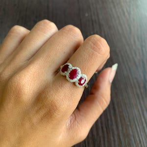 May include: A gold ring featuring three oval-cut, deep red gemstones, each surrounded by a halo of small, clear stones. The ring is set against a dark wooden surface.