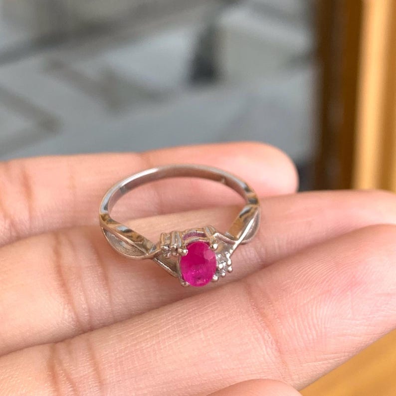 May include: A silver ring with a vivid oval ruby gemstone at its centre. The ring has a delicate design with small accent stones either side of the ruby, adding sparkle. The ring is held in a hand.