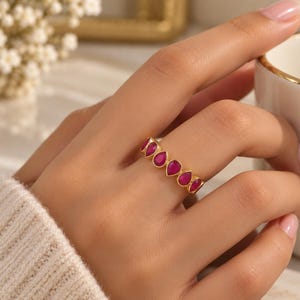 May include: A gold ring featuring a series of vibrant, teardrop-shaped magenta stones set in a continuous row. The band is crafted from a polished gold-tone metal, showcasing a repeating pattern of pear-cut gems that encircle the finger.