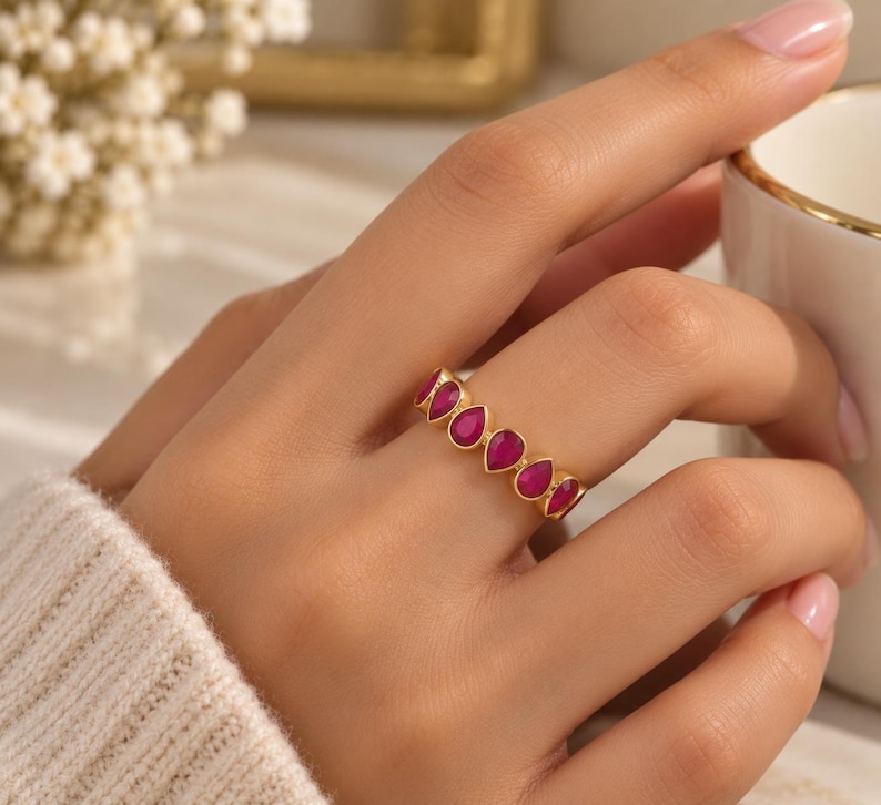 May include: A gold ring featuring a series of vibrant, teardrop-shaped magenta stones set in a continuous row. The band is crafted from a polished gold-tone metal, showcasing a repeating pattern of pear-cut gems that encircle the finger.
